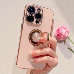 Può includere: Una custodia per telefono rosa con un supporto ad anello color oro. La custodia ha un bordo color oro e una protezione trasparente per l'obiettivo della fotocamera. Il supporto ad anello ha un centro scintillante. Una rosa rosa è sullo sfondo.