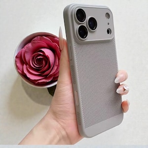 Puede incluir: Una funda gris para teléfono con un diseño perforado, con un módulo de cámara con tres lentes. La funda se sostiene en una mano, con una rosa rosa en un cuenco blanco en el fondo. La funda tiene un acabado liso y mate.