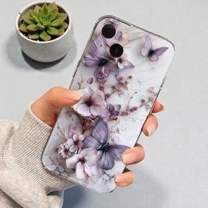 May include: A clear phone case with a 3D floral design. It features lavender butterflies and flowers against a marble-effect background. The case is held in a hand. A small succulent plant in a white pot is visible in the background.
