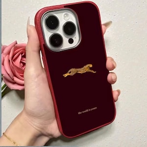 Könnte beinhalten: Burgunderfarbene Handyhülle mit rotem Rand, mit einem goldenen Geparden-Design und dem Text "the world is yours". Die Hülle ist für ein Smartphone mit mehreren Kameralinsen konzipiert.