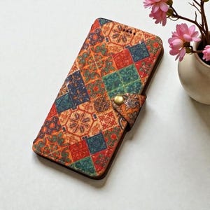 Könnte beinhalten: Eine farbenfrohe Handyhülle mit einem geometrischen Muster in Orange, Rot, Grün und Blau. Die Hülle hat einen Magnetverschluss mit einem goldfarbenen Knopf. Das Design ähnelt einem Patchwork-Quilt.