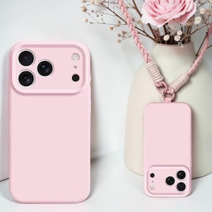 Könnte beinhalten: Zwei hellrosa Handyhüllen, eine mit einem Kameraschutz und eine passende Hülle mit einem geflochtenen rosa Schlüsselband. Die Hüllen werden auf einer weißen Oberfläche mit einer Vase mit Blumen im Hintergrund präsentiert.