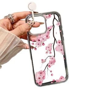 Puede incluir: Funda transparente para teléfono con un diseño de flores de cerezo rosas. La funda tiene un borde transparente y una etiqueta blanca adjunta. El patrón floral incluye flores rosas y ramas marrones.