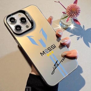 May include: A clear phone case with a silver frame. The case features the name "MESSI" in white letters, with blue and yellow accents. A signature is printed in black. The case is held by a person.