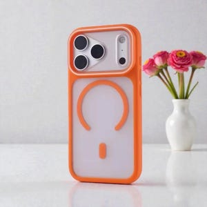 Puede incluir: Funda de teléfono naranja con parte trasera transparente y borde blanco alrededor de las lentes de la cámara. La funda tiene un diseño circular en la parte trasera. Un pequeño jarrón con flores rosas en el fondo.