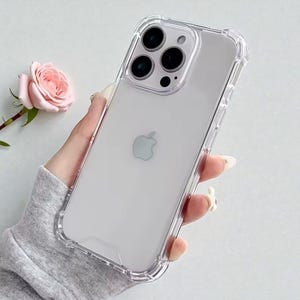 Könnte beinhalten: Transparente Schutzhülle für ein weißes iPhone. Die Hülle hat ein klares Design und einen erhöhten Rand. Das Telefon verfügt über eine Dreifach-Kamera und das Apple-Logo. Die Hülle schützt das Telefon vor Kratzern und Beschädigungen.
