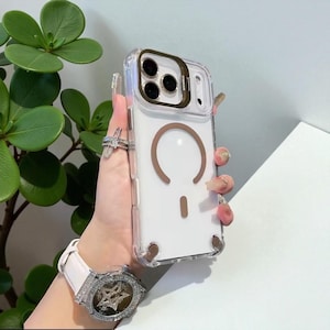Puede incluir: Funda transparente para teléfono con diseño de carga magnética marrón. La funda tiene un anillo de cámara dorado y detalles en marrón. La funda se sostiene frente a una planta verde.