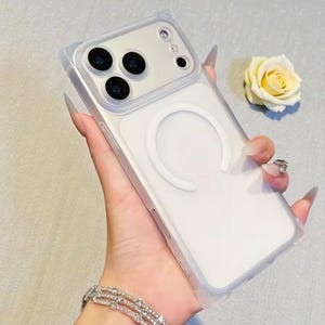 May include: Clear phone case with a white magnetic ring and a transparent bumper. The case is designed to fit a smartphone with multiple camera lenses. A yellow rose is in the background.