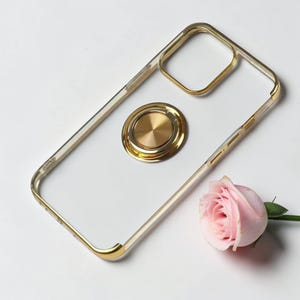 May include: Transparent phone case with a gold-coloured frame and a gold ring holder. The case is shown on a white surface with a pink rose.