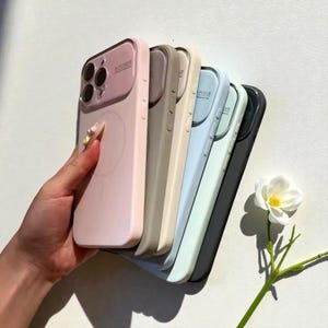 Könnte beinhalten: Eine Reihe farbiger Handyhüllen, gehalten von einer Hand. Die Hüllen sind in den Farbtönen Rosa, Beige, Hellblau, Mintgrün und Schwarz gehalten. Jede Hülle hat einen Kameraschutz mit dem Text "AUTO FOCUS".