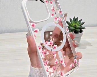 May include: Clear phone case with a white border and a floral design featuring pink blossoms. The case has a circular magnetic charging area and is designed to fit a smartphone.
