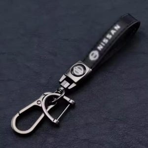 May include: Black leather keychain with a silver-tone metal clasp and hardware. The keychain features the Nissan logo and brand name in white lettering. The design is sleek and modern.