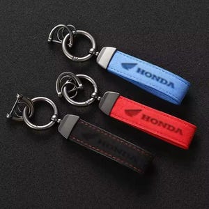Könnte beinhalten: Drei Schlüsselanhänger mit Lederriemen in Blau, Rot und Schwarz. Jeder Schlüsselanhänger hat einen silbernen Metallring und einen Riemen mit dem Aufdruck "HONDA". Die blauen und roten Riemen haben ein Honda-Logo.
