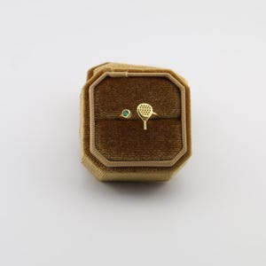 May include: A gold ring with a tennis racket design and a small green gemstone, displayed in a brown velvet ring box. The ring box is octagonal and has a gold trim.