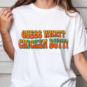 Pode incluir: T-shirt branco com a frase "GUESS WHAT? CHICKEN BUTT!" em laranja, amarelo e turquesa com um estilo retro. A t-shirt é de estilo casual, adequada para o uso diário.