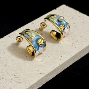May include: Gold-plated hoop earrings with a sea-themed design. The earrings have blue, white, and green enamel detailing, with seashell and pearl embellishments. They are presented on a textured, light-coloured surface.