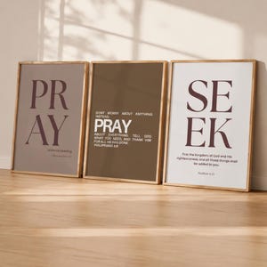 May include: Three framed wall art prints with light wood frames. The left print is mauve with the word PRAY and text 1 Thessalonians 5:17. The center print is brown with the text PRAY and Philippians 4:6. The right print is white with SEEK and Matthew 6:33.
