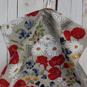 Op de afbeelding: Een beige theedoek met een bloemenpatroon met rode klaprozen en witte madeliefjes. Het patroon bevat ook tarwehalmen en andere kleine bloemdetails.