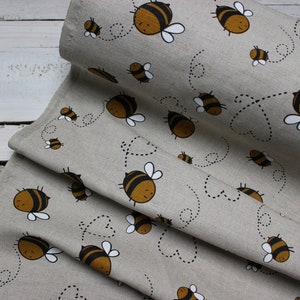 Peut inclure: Tissu beige avec un motif répété d'abeilles de dessin animé à rayures marron et noires, avec des ailes blanches. Les abeilles sont entourées de trajectoires de vol pointillées. Le tissu est plié.