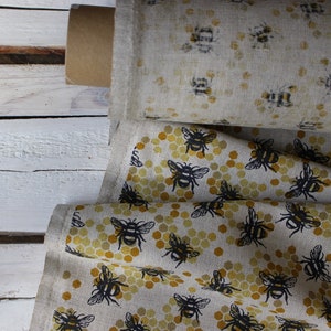 Peut inclure: Deux rouleaux de tissu avec un motif d'abeilles et de nids d'abeilles. Le rouleau du haut a un fond blanc avec des abeilles noires. Le rouleau du bas a un fond beige avec des abeilles noires et des nids d'abeilles jaunes.