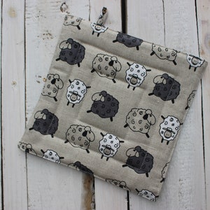 Peut inclure: Un dessous de plat carré beige avec un motif répété de moutons de dessin animé noirs, blancs et gris. Le dessous de plat a une boucle pour l'accrocher et est fait de tissu matelassé. Le fond est une surface en bois blanc.