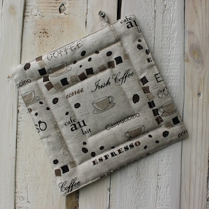 May include: Square beige pot holder with a coffee theme. The pot holder features coffee cups, coffee beans, and text including "Coffee", "Espresso", "Irish Coffee", and "Cappuccino". The pot holder has a loop for hanging.
