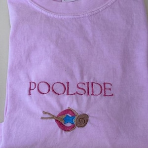 May include: A light pink t-shirt with the word "POOLSIDE" embroidered in a darker pink thread. Below the text is an embroidered design of a person floating in a pool with a blue star and a brown hat.