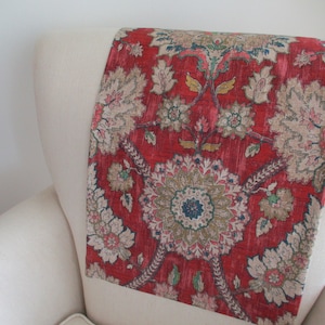 May include: A red and beige floral patterned textile draped over the back of a white armchair. The textile features a central medallion design with intricate floral and leaf motifs in shades of beige, green, and blue. The background is a deep red.