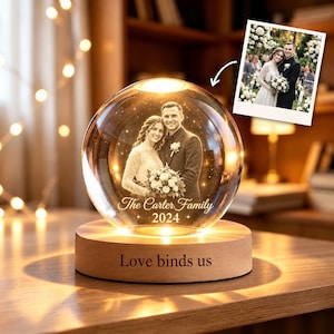 May include: A clear glass sphere featuring a laser-etched image of a couple, with the text "The Carter Family 2024" engraved inside. The sphere rests on a round wooden base with the engraved text "Love binds us" and includes an inset photo.