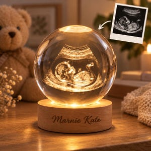 May include: A clear glass sphere lamp featuring a 3D laser-engraved ultrasound image of a fetus inside. The sphere rests on a round, light-colored wooden base with the name Marnie Kate engraved in a cursive script on the front surface.
