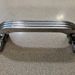 Puede incluir: Un tirador cromado para armario. El tirador tiene un diseño curvo y rectangular con ranuras horizontales. Las placas de montaje tienen detalles decorativos. Adecuado para armarios, cajones o muebles.