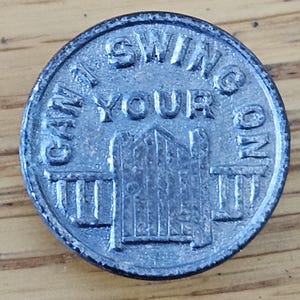 May include: A circular, silver-colored metal token with the words "CAN I SWING ON YOUR" around the top and left side. The words "ON" are on the right side. A gate design is in the center.