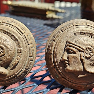 May include: Two bronze-colored cabinet knobs with a classical design. Each knob features a profile of a person wearing a helmet, surrounded by a decorative border. The knobs are round and appear to be made of metal.