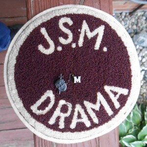 May include: A round, maroon-colored felt item with the words "J.S.M. DRAMA" in white lettering. A small silver pin with a skull design and the letter "M" are attached to the felt. The item has a cream-colored border.