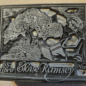 May include: A rectangular, gray metal bookplate with a detailed engraved design. The image features a woman reading under a tree, with the text "Ex Libris Eloise Ramsey" and the number "No. 6". The style is reminiscent of vintage book illustrations.