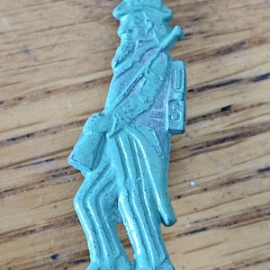 May include: A vintage, turquoise-colored metal pin depicting Uncle Sam. The pin features Uncle Sam in his iconic top hat and suit, carrying a rolled-up document. The letters "US" are visible on the back of the figure.