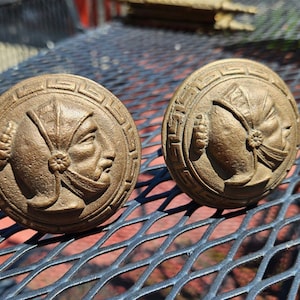 May include: Two bronze-colored, circular cabinet knobs. Each knob features a detailed relief of a profile of a person wearing a head covering and a decorative border. The knobs are displayed on a dark metal surface.