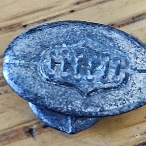 May include: A weathered, oval-shaped metal object with the embossed text "HHH" within a heart-shaped design. The object has a textured, aged appearance, and is set against a wooden surface.