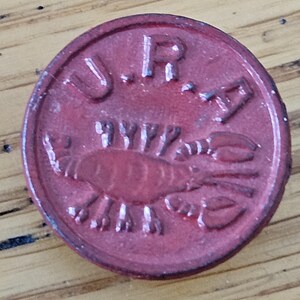 May include: A round, red button with the embossed text "U.R.A." and a detailed image of a lobster. The button has a slightly worn appearance, suggesting age or use. The lobster is centered, with its claws and body clearly defined.