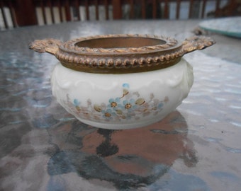 May include: A white, round, antique bowl with a bronze rim and handles. The bowl is decorated with a floral pattern in blue and brown. The rim and handles have an ornate, textured design.