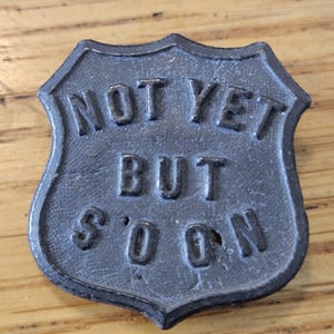 May include: A dark gray metal shield-shaped plaque with the words "NOT YET BUT SOON" embossed on it. The plaque has a slightly weathered appearance and is set against a wooden surface.