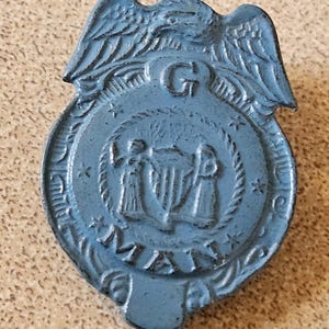 May include: A blue metal badge with an eagle at the top and the word "MA" at the bottom. The badge has a circular center with two figures and a shield. The letter "G" is at the top center.