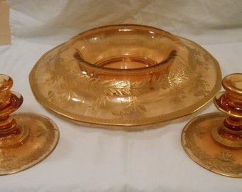 May include: A set of amber-colored glass serving pieces, including a large bowl and two candle holders. The pieces feature a gold leaf design around the edges. The bowl has a wide rim and a central depression.