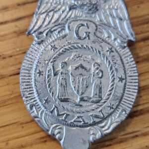 May include: A silver-colored metal badge with an eagle at the top and a circular emblem in the center. The emblem features two figures flanking a shield, with the word "MAN" at the bottom. The badge has a vintage, possibly antique, appearance.