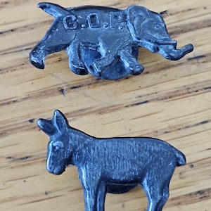 May include: Two dark gray metal pins. The top pin is an elephant with the letters "G.O.P." on its side. The bottom pin is a donkey. Both pins are on a light brown wooden surface.