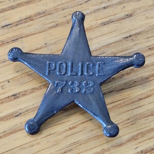 May include: A vintage-style, five-pointed star-shaped police badge. The badge is dark gray with the word "POLICE" and the number "732" embossed on its surface. The badge has rounded tips and is set against a wooden background.