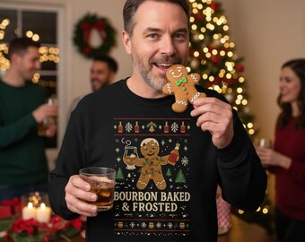 Op de afbeelding: Een zwarte sweatshirt met een feestelijk ontwerp met een peperkoekmannetje met een whiskyglas en een fles. De tekst "BOURBON BAKED & FROSTED" staat eronder. De persoon houdt een peperkoek en een glas whisky vast.