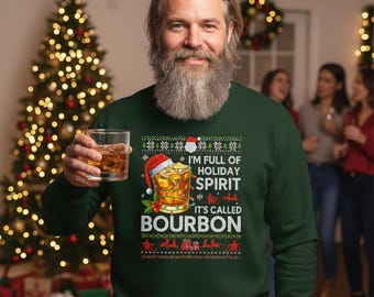 Op de afbeelding: Een donkergroene sweatshirt met een vakantie-thema ontwerp. De afbeelding bevat een cocktailglas met een kerstmuts, hulst en de tekst "I'M FULL OF HOLIDAY SPIRIT IT'S CALLED BOURBON". Een man met een baard houdt een glas amberkleurige vloeistof vast.