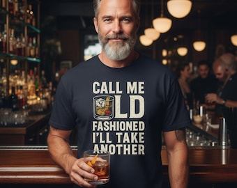 Pode incluir: Camiseta azul marinho com o texto "CALL ME OLD FASHIONED I'LL TAKE ANOTHER" e um gráfico de um coquetel Old Fashioned. A pessoa na imagem está segurando um copo do mesmo coquetel.