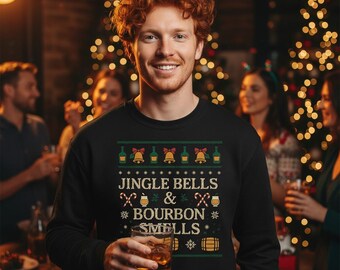 Op de afbeelding: Zwart sweatshirt met een feestelijk ontwerp met flessen, bellen en de tekst "Jingle Bells & Bourbon Smells". Het ontwerp is in groen, goud en wit. De persoon op de foto houdt een glas amberkleurige vloeistof vast.